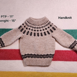 May include: Hand-knitted beige sweater with a high collar and long sleeves. The sweater features a dark brown geometric pattern around the collar, cuffs, and hem. The PTP is 28 cm, and the length is 38 cm. The word "Handknit" is visible.