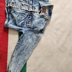 May include: A blue denim jacket with a stone wash effect. The jacket features multiple pockets, button closures, and a classic collar. The sleeves are long, and the fabric has a faded, textured appearance. The jacket is folded on a striped blanket.