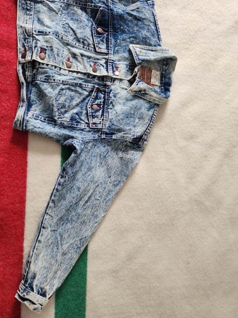 May include: A blue denim jacket with a stone wash effect. The jacket features multiple pockets, button closures, and a classic collar. The sleeves are long, and the fabric has a faded, textured appearance. The jacket is folded on a striped blanket.