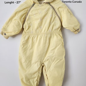 Peut inclure: Combinaison de neige jaune pâle pour bébé, avec un col montant et des manches longues. La combinaison a une fermeture éclair et des détails brodés décoratifs. Le texte "Pixie Togs Toronto Canada" est visible. Mesures: 28 cm de largeur et 69 cm de longueur.