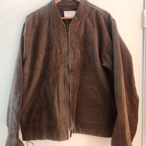 Peut inclure: Veste de bombardier marron avec une fermeture &eacute;clair int&eacute;grale, deux poches avant et des poignets c&ocirc;tel&eacute;s. La veste est suspendue &agrave; un cintre en bois sur fond blanc. Le mat&eacute;riau de la veste semble &ecirc;tre du daim ou un tissu similaire.