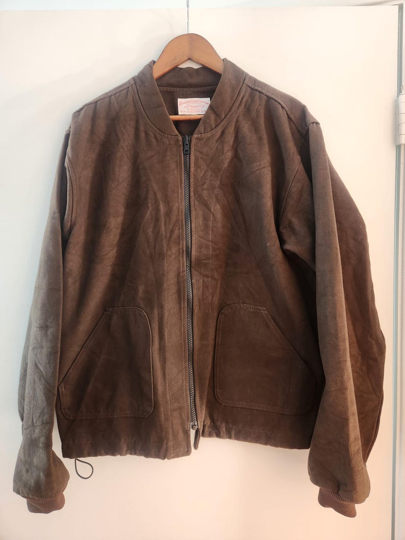 Peut inclure: Veste de bombardier marron avec une fermeture &eacute;clair int&eacute;grale, deux poches avant et des poignets c&ocirc;tel&eacute;s. La veste est suspendue &agrave; un cintre en bois sur fond blanc. Le mat&eacute;riau de la veste semble &ecirc;tre du daim ou un tissu similaire.