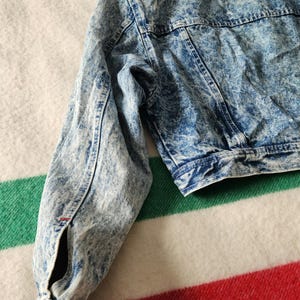 May include: A cropped acid wash denim jacket in shades of blue and white. The jacket features a classic collar, button closures, and long sleeves. The fabric has a textured, faded appearance, with visible stitching details.