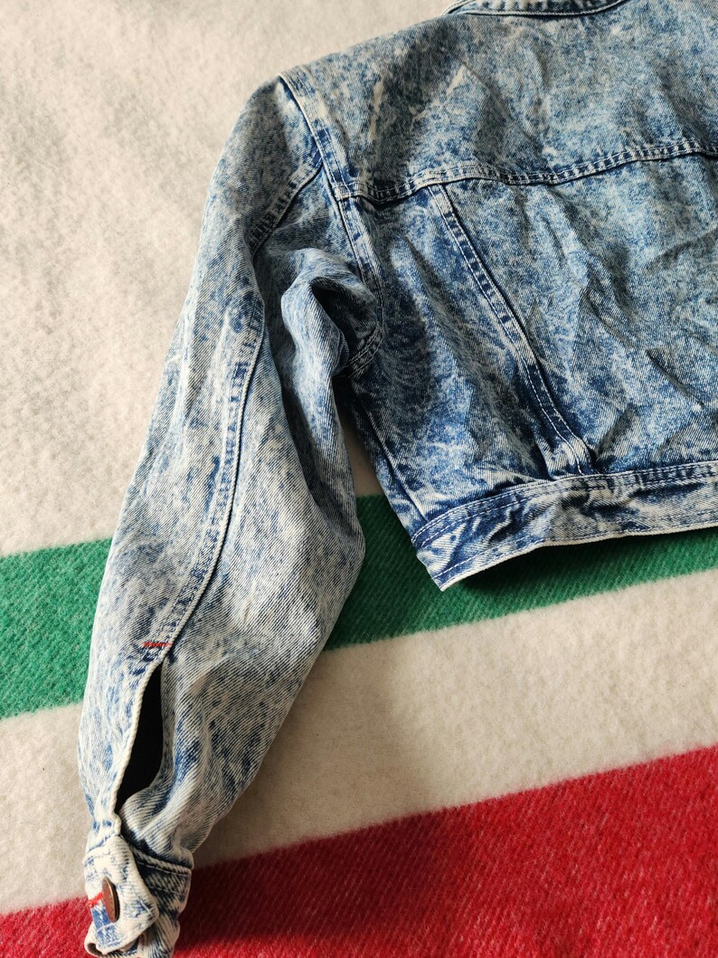 May include: A cropped acid wash denim jacket in shades of blue and white. The jacket features a classic collar, button closures, and long sleeves. The fabric has a textured, faded appearance, with visible stitching details.
