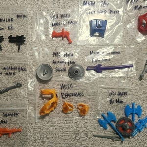 May include: A selection of vintage MOTU action figure accessories, including weapons, shields, and character-specific parts. The items are in small plastic bags, with handwritten labels. Colours include black, orange, blue, and red.