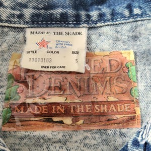 May include: Close-up of a denim jacket with a faded blue wash. The tag reads "MADE IN THE SHADE" and includes the text "CRAFTED WITH PRIDE IN USA." The tag also shows the size "S".