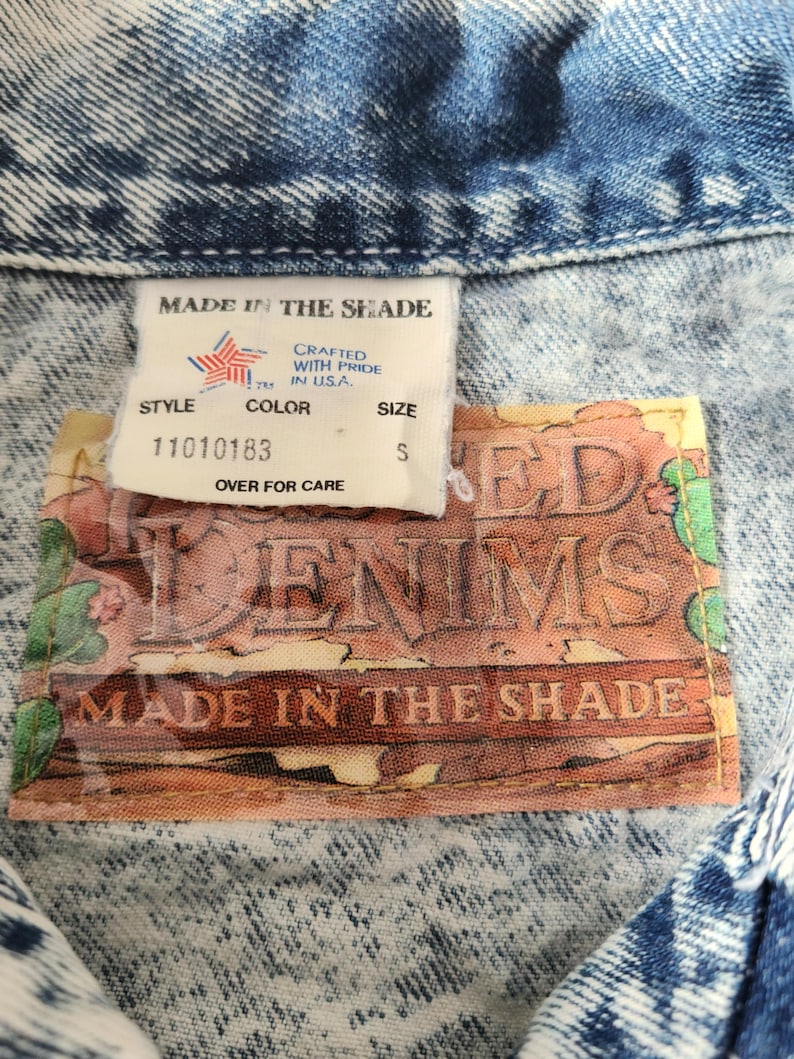 May include: Close-up of a denim jacket with a faded blue wash. The tag reads "MADE IN THE SHADE" and includes the text "CRAFTED WITH PRIDE IN USA." The tag also shows the size "S".