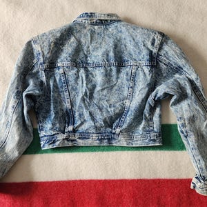 May include: A vintage acid wash denim jacket, with a classic collar, button-up front, and long sleeves. The jacket has a light blue and white mottled pattern, with two chest pockets and a tailored fit. The jacket is approximately 51 cm long.