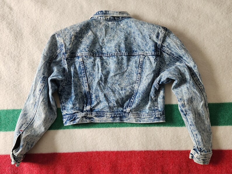 May include: A vintage acid wash denim jacket, with a classic collar, button-up front, and long sleeves. The jacket has a light blue and white mottled pattern, with two chest pockets and a tailored fit. The jacket is approximately 51 cm long.