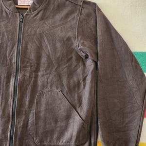 Peut inclure: Veste de bombardier marron avec une fermeture &eacute;clair pleine longueur et une seule poche. La veste a un poignet et un ourlet c&ocirc;tel&eacute;s. La veste est suspendue &agrave; un cintre en bois.