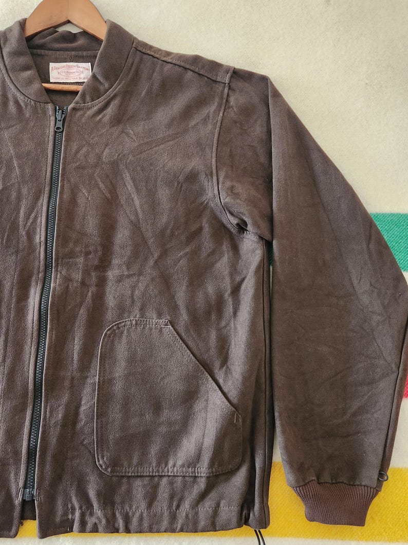 Peut inclure: Veste de bombardier marron avec une fermeture &eacute;clair pleine longueur et une seule poche. La veste a un poignet et un ourlet c&ocirc;tel&eacute;s. La veste est suspendue &agrave; un cintre en bois.