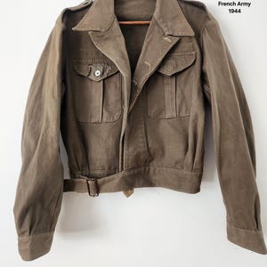 May include: Olive green bomber jacket from the French Army, dated 1944. Features a belted waist, two front pockets, and a collared neckline. Measurements: PTP - 20", Lenght - 21", Shoulder to cuff - 23".