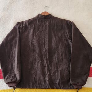 Peut inclure: Veste marron fonc&eacute; avec un col montant et des manches longues. La veste a un cordon de serrage en bas et est suspendue &agrave; un cintre en bois. Le tissu semble &ecirc;tre une mati&egrave;re douce, peut-&ecirc;tre semblable &agrave; du daim. L'arri&egrave;re-plan est une couverture ray&eacute;e.