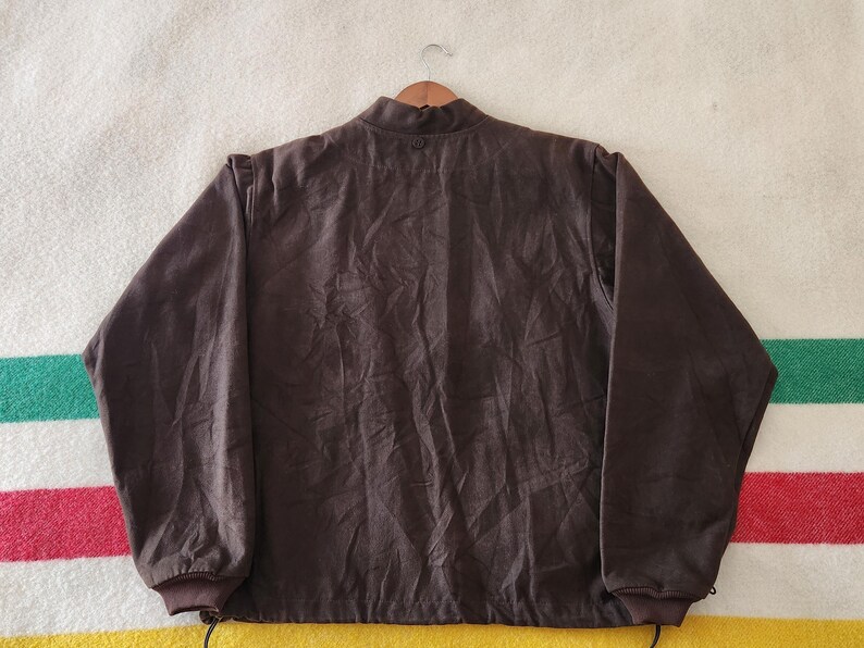 Peut inclure: Veste marron fonc&eacute; avec un col montant et des manches longues. La veste a un cordon de serrage en bas et est suspendue &agrave; un cintre en bois. Le tissu semble &ecirc;tre une mati&egrave;re douce, peut-&ecirc;tre semblable &agrave; du daim. L'arri&egrave;re-plan est une couverture ray&eacute;e.