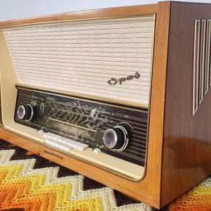 May include: A retro wooden radio featuring a cream speaker grille and a dark control panel with dials and buttons. The word "Opus" is visible on the front. The radio is housed in a wooden cabinet and sits on a patterned blanket.