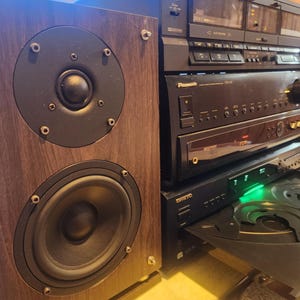 May include: A vintage stereo system featuring a wooden speaker cabinet with two black speakers. The system includes a cassette deck, a receiver, and a CD player, all in black.