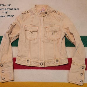 以下が含まれることがあります： オレンジ色のステッチが施されたベージュのジャケット。フロントポケットが2つ、ボタンのディテールがあります。スタンドカラーとボタン付きの袖口。サイズ：PTP - 38 cm、肩からフロントヘム - 48 cm、袖 - 60 cm。