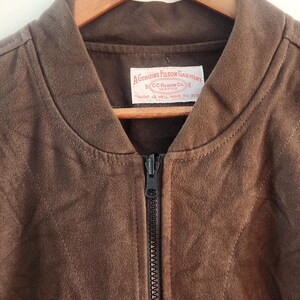 Peut inclure: Veste de bombardier en daim marron avec une fermeture &eacute;clair sur toute la longueur. La veste est suspendue &agrave; un cintre en bois. Une &eacute;tiquette &agrave; l'int&eacute;rieur du col indique "A GENUINE FILSON GARMENT" et "MIGHT AS WELL HAVE THE BEST".