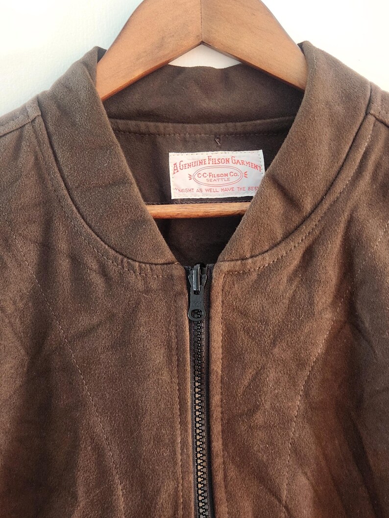 Peut inclure: Veste de bombardier en daim marron avec une fermeture &eacute;clair sur toute la longueur. La veste est suspendue &agrave; un cintre en bois. Une &eacute;tiquette &agrave; l'int&eacute;rieur du col indique "A GENUINE FILSON GARMENT" et "MIGHT AS WELL HAVE THE BEST".