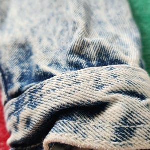 May include: Close-up of a folded denim cuff. The fabric is light blue with a faded, acid-wash effect. The cuff's edge is a lighter shade of blue, showing the fabric's texture and weave. The overall look is casual and vintage.