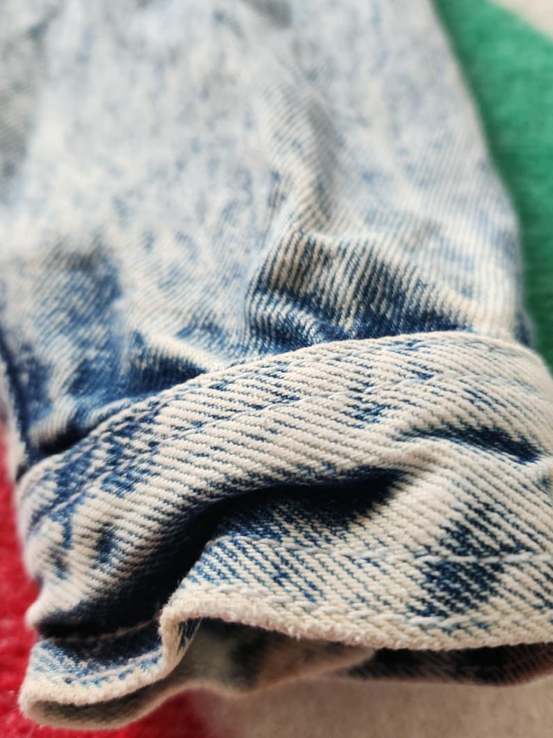 May include: Close-up of a folded denim cuff. The fabric is light blue with a faded, acid-wash effect. The cuff's edge is a lighter shade of blue, showing the fabric's texture and weave. The overall look is casual and vintage.