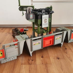 Pode incluir: Um conjunto de brinquedo G.I. Joe com um centro de comando de vários níveis, uma torre verde alta, um braço de guindaste e uma plataforma central. A base inclui seções de parede modulares cinzentas com painéis laranja e faixas amarelas.