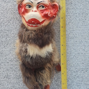 May include: Vintage monkey doll with a painted face and fur body. The face is painted with red cheeks, lips, and ears. The doll measures approximately 10 inches in length. The fur is brown and tan. The doll has wooden feet and hands.