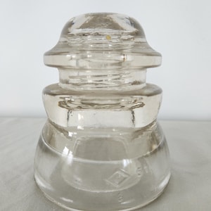 Peut inclure: Un isolateur électrique en verre transparent présentant un profil arrondi à plusieurs niveaux avec une surface lisse et translucide. La base de l'objet affiche un logo en forme de losange moulé avec le chiffre 51 gravé dans le verre.