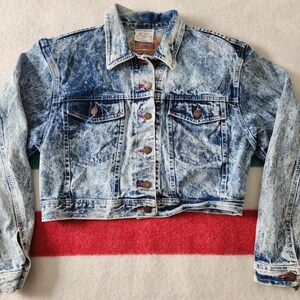 May include: A vintage acid wash denim jacket. It has a classic collar, button-down front, and two chest pockets. The jacket features a faded blue and white pattern, with brown buttons and red stitching accents. A timeless piece for casual wear.