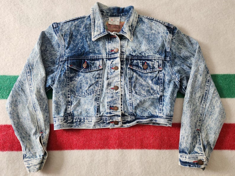 May include: A vintage acid wash denim jacket. It has a classic collar, button-down front, and two chest pockets. The jacket features a faded blue and white pattern, with brown buttons and red stitching accents. A timeless piece for casual wear.