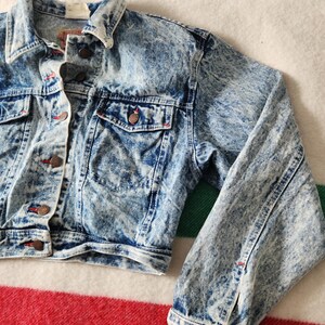May include: Light blue acid wash denim jacket with button fastenings, two chest pockets, and side pockets. The jacket features a classic collar and buttoned cuffs. The denim has a white, marbled effect. Red stitching accents the pockets and seams.