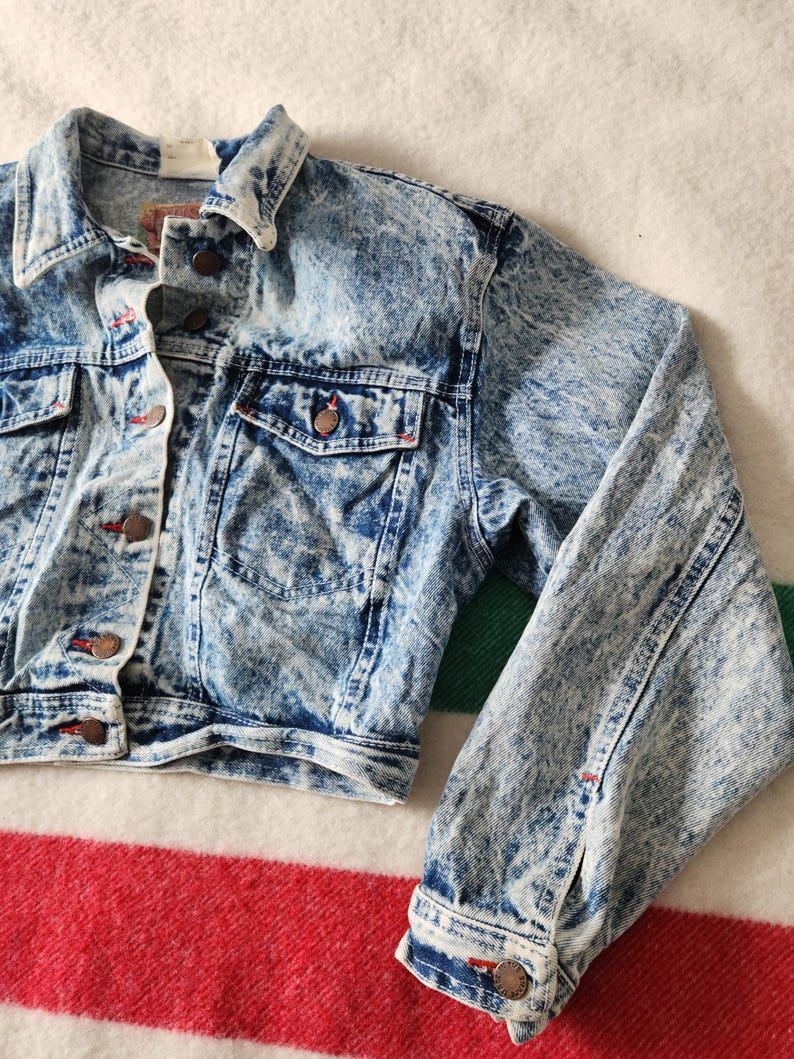 May include: Light blue acid wash denim jacket with button fastenings, two chest pockets, and side pockets. The jacket features a classic collar and buttoned cuffs. The denim has a white, marbled effect. Red stitching accents the pockets and seams.