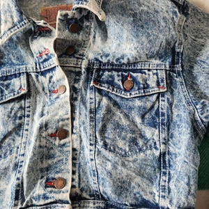 May include: A close-up of a vintage acid wash denim jacket. The jacket has a classic collar, button-down front, and two chest pockets. Red stitching accents the pockets and buttonholes. The denim has a blue and white marbled effect.