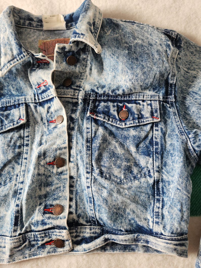 May include: A close-up of a vintage acid wash denim jacket. The jacket has a classic collar, button-down front, and two chest pockets. Red stitching accents the pockets and buttonholes. The denim has a blue and white marbled effect.