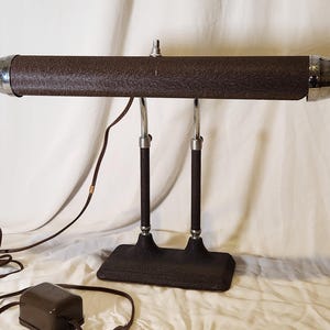 Puede incluir: Una lámpara de escritorio vintage con una pantalla marrón texturizada y detalles cromados. La lámpara tiene una base rectangular y dos soportes curvos. El cable de alimentación y el transformador son visibles. Una lámpara clásica.