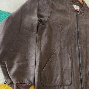 Peut inclure: Veste de bombardier marron avec une fermeture &eacute;clair int&eacute;grale, deux poches avant et des poignets c&ocirc;tel&eacute;s. La veste est suspendue &agrave; un cintre en bois. Le tissu semble &ecirc;tre une mati&egrave;re douce semblable &agrave; du daim.