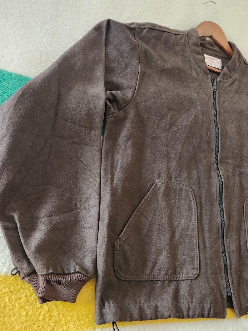 Peut inclure: Veste de bombardier marron avec une fermeture &eacute;clair int&eacute;grale, deux poches avant et des poignets c&ocirc;tel&eacute;s. La veste est suspendue &agrave; un cintre en bois. Le tissu semble &ecirc;tre une mati&egrave;re douce semblable &agrave; du daim.