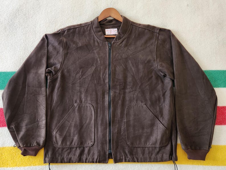 Peut inclure: Veste de bombardier marron avec une fermeture &eacute;clair sur toute la longueur et deux poches avant. La veste a un col et des poignets c&ocirc;tel&eacute;s. La veste est suspendue &agrave; un cintre en bois.