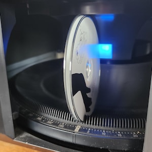 Könnte beinhalten: Ein schwarzer CD-Player mit einer Disc im Lademechanismus. Die Disc ist weiß mit einem schwarzen Design. Ein blaues Licht scheint auf die Disc.