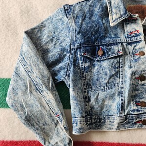 May include: A vintage acid wash denim jacket in a light blue and white marbled pattern. It has a classic collar, button-up front, and two chest pockets. The jacket features red stitching and copper-coloured buttons. The sleeves are cuffed at the wrists.