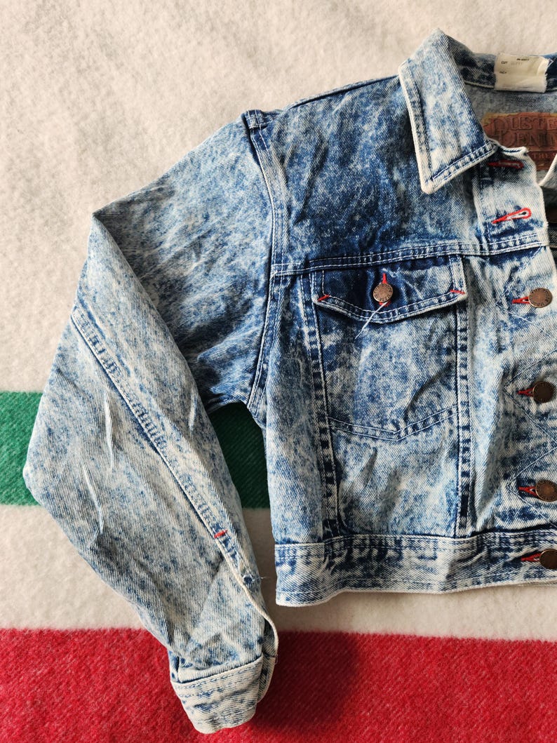 May include: A vintage acid wash denim jacket in a light blue and white marbled pattern. It has a classic collar, button-up front, and two chest pockets. The jacket features red stitching and copper-coloured buttons. The sleeves are cuffed at the wrists.