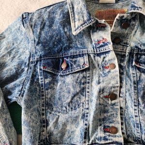 May include: Acid wash denim jacket with a button-up front, two chest pockets, and a classic collar. The jacket features red stitching details and copper-coloured buttons. The label reads "Dusted Denim".