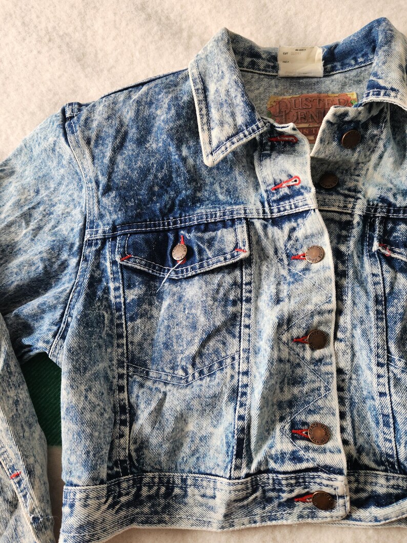 May include: Acid wash denim jacket with a button-up front, two chest pockets, and a classic collar. The jacket features red stitching details and copper-coloured buttons. The label reads "Dusted Denim".