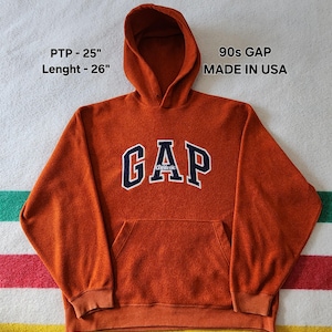 Peut inclure: Un sweat à capuche GAP orange des années 90 avec une capuche et une poche avant. Le mot "GAP" est sur la poitrine en bleu marine avec un contour blanc, et le mot "Athletic" est en dessous. Fabriqué aux États-Unis. PTP - 63.5 cm, Longueur - 66 cm.