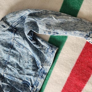 May include: A light blue acid wash denim jacket with a classic collar and button fastenings. The jacket is laid on a striped blanket with red, white, and green stripes. The jacket's sleeves are visible.