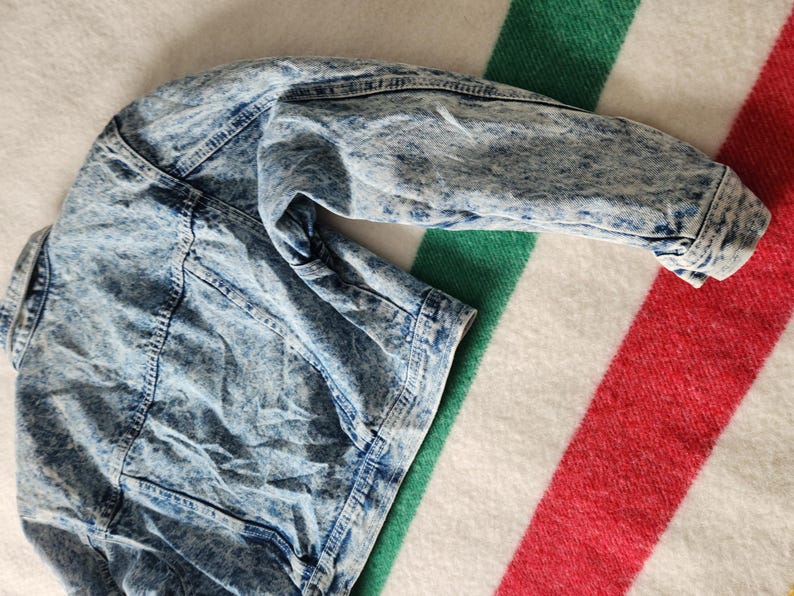 May include: A light blue acid wash denim jacket with a classic collar and button fastenings. The jacket is laid on a striped blanket with red, white, and green stripes. The jacket's sleeves are visible.