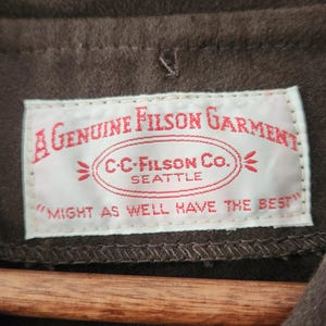 Peut inclure: Gros plan sur un v&ecirc;tement marron avec une &eacute;tiquette blanche. L'&eacute;tiquette indique "A Genuine Filson Garment" avec "C.C. Filson Co. Seattle" et la phrase "Might as well have the best."