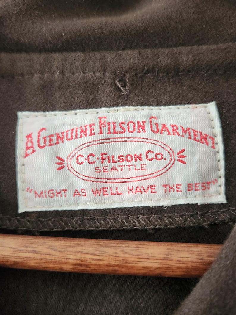 Peut inclure: Gros plan sur un v&ecirc;tement marron avec une &eacute;tiquette blanche. L'&eacute;tiquette indique "A Genuine Filson Garment" avec "C.C. Filson Co. Seattle" et la phrase "Might as well have the best."