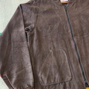 Peut inclure: Veste de bombardier marron avec une fermeture &eacute;clair noire sur toute la longueur et deux poches avant. La veste a un poignet c&ocirc;tel&eacute; et une petite &eacute;tiquette pr&egrave;s du col. La veste est suspendue &agrave; un cintre en bois.