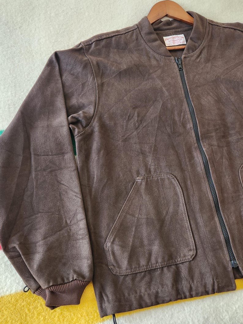 Peut inclure: Veste de bombardier marron avec une fermeture &eacute;clair noire sur toute la longueur et deux poches avant. La veste a un poignet c&ocirc;tel&eacute; et une petite &eacute;tiquette pr&egrave;s du col. La veste est suspendue &agrave; un cintre en bois.